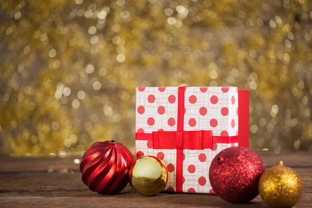 Festive scene featuring a wrapped gift box with red polka dots and a red ribbon, accompanied by red and gold Christmas baubles on a wooden table. Ideal for holiday-themed promotions, greeting cards, festive advertisements, and seasonal blog posts.