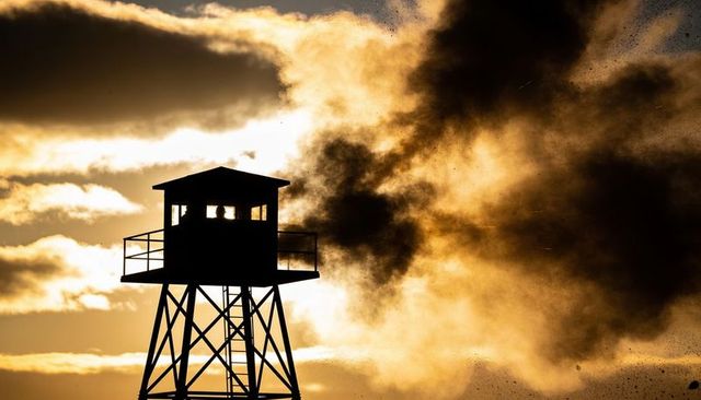 Silhouetted watchtower cabin with observers at golden sunset and dramatic smoke plume