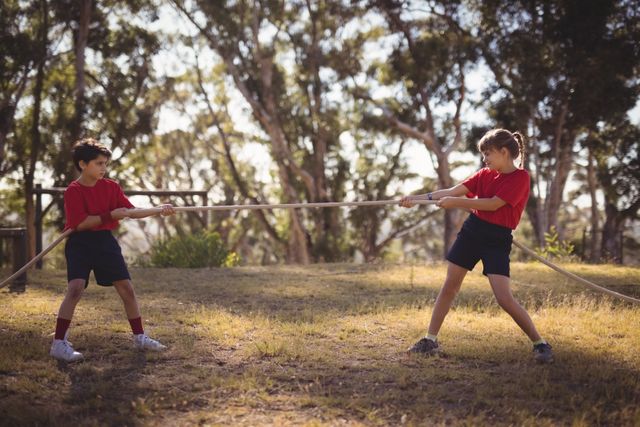 Two children are engaged in a tug of war during a boot camp obstacle course. They are outdoors, surrounded by trees, and appear focused and determined. This image can be used to illustrate teamwork, childhood activities, outdoor sports, and summer camp experiences.