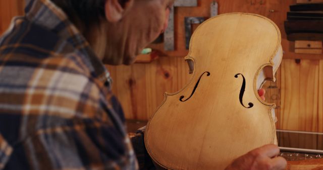 Senior craftsman carefully inspecting a hand-crafted violin in a workshop. Ideal for articles on traditional craftsmanship, musical instrument making, woodworking artistry, and features on aging artisans. Can be used to illustrate dedication and skilled work in niche professions.