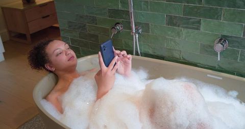 African american woman relaxing in bubble bath using smartphone in modern green bathroom