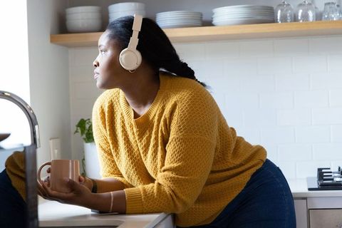 African american woman leaning at kitchen sink wearing headphones and holding coffee mug
