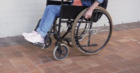 Young woman in wheelchair placing hand on rim on brick courtyard, denim and pink sneakers