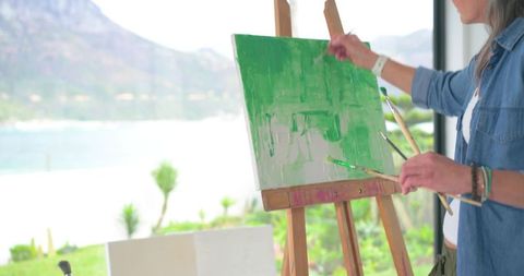 Woman painting green abstract on canvas at sunlit lakefront studio with mountain view