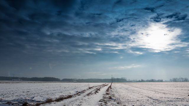 Scenic snow covered rural field under a dramatic cloudy sky. Ideal image for use in seasonal marketing, holiday cards, nature blogs, environmental articles, and travel advertisements focusing on serene countryside landscapes.