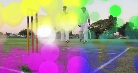 Two cricketers walking on sunset pitch carrying bats with colorful bokeh overlay