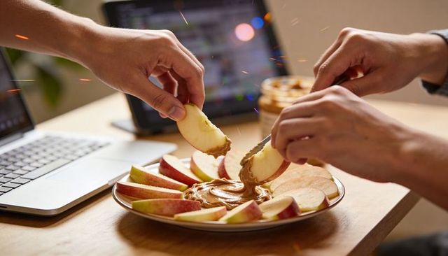 Hands dipping apple slices into creamy peanut butter on desk near laptop for snack casual