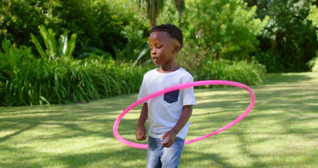 African American boy playing with a pink hula hoop in a lush green park. Great for themes related to childhood activities, outdoor fun, healthy lifestyle, summer, and exercise.