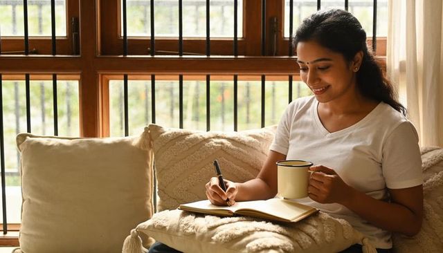 Sunlit woman journaling with mug on cozy sofa by wooden window, mindful home lifestyle