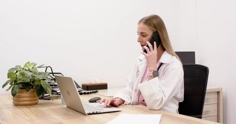 Female doctor calling patient while typing on laptop in minimalist modern clinic office