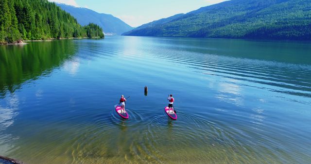 Two people are paddleboarding on a serene lake surrounded by mountains, with copy space. Engaging in water sports, they enjoy the tranquility and natural beauty of the landscape.