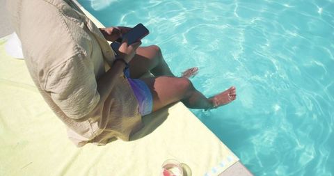 African american man sitting poolside using smartphone dipping feet in turquoise water