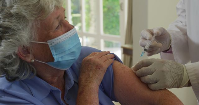 An elderly woman is receiving a COVID-19 vaccination from a healthcare worker. She is wearing a blue shirt and a protective face mask, and the healthcare worker is wearing gloves. The vaccination is being administered inside a bright room. This image can be used in articles and campaigns related to vaccination, healthcare, pandemic safety measures, elderly care, and public health initiatives.