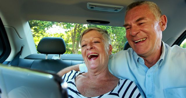 Elderly couple seated in backseat of a car using a digital device for video calling. They are laughing and enjoying the moment, showcasing technology's ability to connect people of all ages. Ideal for use in content related to senior living, technology adoption among the elderly, family bonding, and travel.