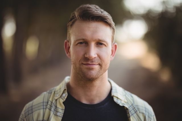 Close-up portrait of a handsome young man standing outdoors at an olive farm. He is wearing a casual plaid shirt and smiling confidently. The natural light and rural background create a serene and approachable atmosphere. Ideal for use in lifestyle, outdoor activities, and agricultural themes.