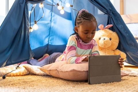 Young african american girl using tablet inside cozy blue tent with teddy bear