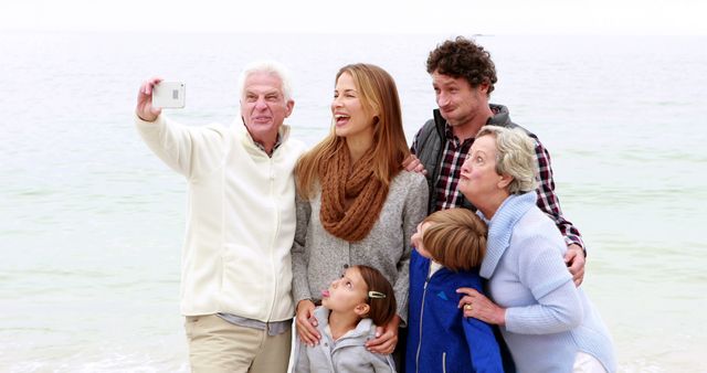 This image shows a multigenerational family taking a selfie at the beach. They are standing close together, smiling and appearing very happy. The background reveals a peaceful ocean. This image can be used for family-oriented advertisements, travel brochures, articles on family bonding, or blogs discussing beach vacations.