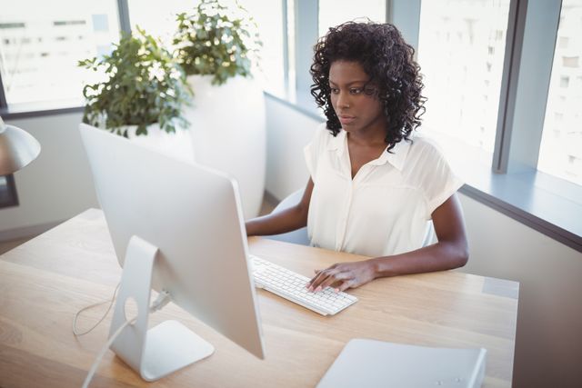 This image shows a professional woman working at her desk in a modern office environment. She is focused on her computer, indicating productivity and dedication. This image can be used for business, corporate, and technology-related content, such as articles on workplace efficiency, office environments, or professional development.