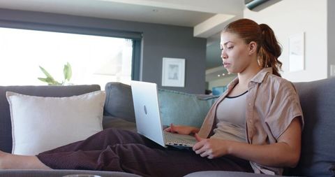 Woman working from home on laptop in loungewear on modern sofa, relaxed remote work