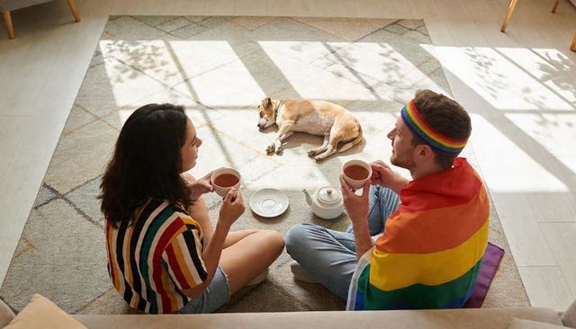 Couple sharing tea on sunlit rug with rainbow flag and sleeping dog, cozy lgbtq morning moment
