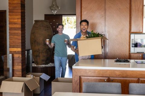 Diverse roommates carrying moving boxes into modern wood kitchen holding plant and coffee