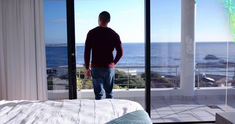 Contemplative person standing by sliding glass door overlooking ocean from coastal bedroom