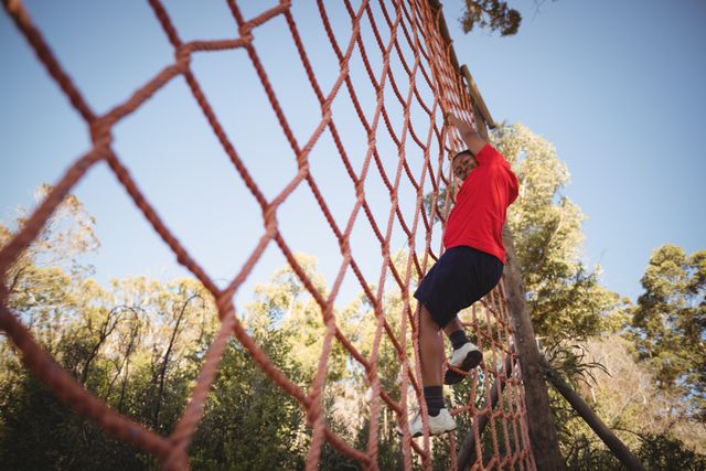 Boy climbing a net during an obstacle course in a boot camp. He is smiling and appears determined. The background shows trees and a clear sky, indicating an outdoor setting. This image can be used for promoting outdoor activities, fitness programs, summer camps, and adventure challenges.