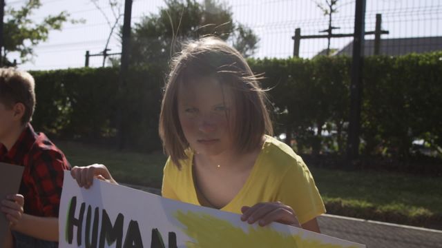 Caucasian girl looking directly at the camera while holding a sign with environmental slogans. Other children seen in background participating in protest. Ideal for materials on environmental education, children’s activism, school awareness campaigns, and eco-friendly initiatives.