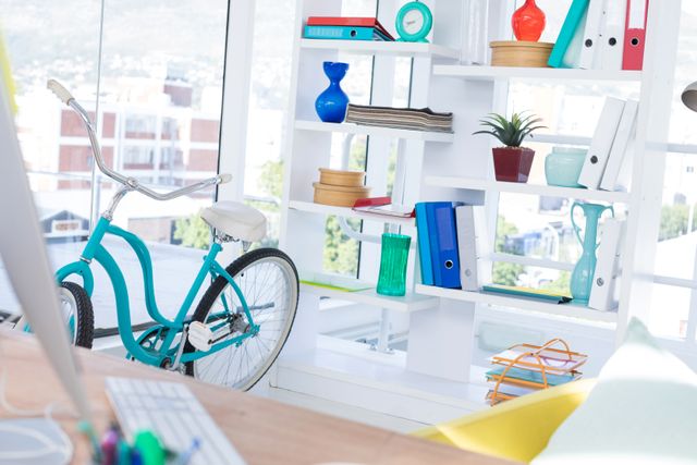 Bright and modern office featuring a bicycle and a well-organized shelf. The workspace includes a computer on a desk, various office supplies, and decor items such as an indoor plant and colorful binders. Ideal for illustrating contemporary work environments, creative workspaces, and urban office settings.