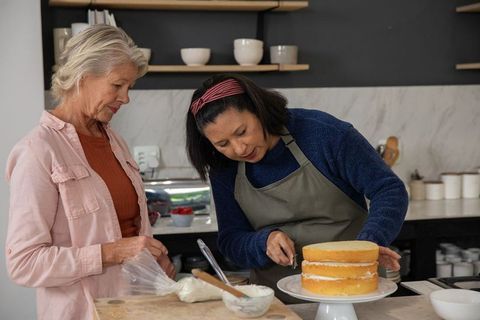 Senior women decorating layered sponge cake together in cozy home kitchen