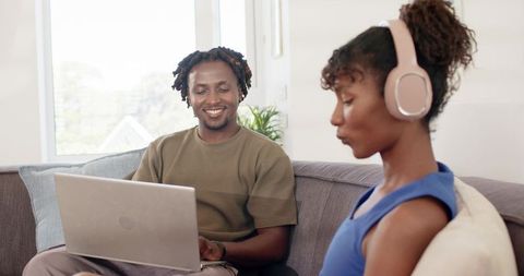 African american couple using laptop at home while partner wearing pink headphones and smiling