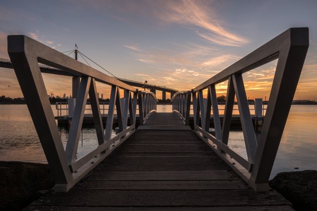 Wooden walkway leading to dock with waterfront bridge at sunset depicted. Features calm water and a vibrant sky. Useful for backgrounds, travel blogs, architecture presentations, and landscape photography collections.