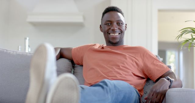 Young African American man smiling while sitting on a comfortable couch in a modern living room. Ideal for articles on relaxation, home comfort, casual lifestyle, happiness, or interior design themes.