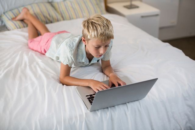 Young boy lying on bed using laptop in a cozy bedroom. Ideal for illustrating concepts related to childhood education, technology use among children, home learning environments, and relaxed leisure activities. Suitable for articles, blogs, and advertisements focusing on kids' tech engagement, online learning, and home lifestyle.