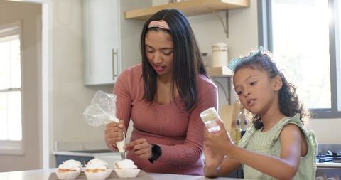 Diverse mother and daughter decorating cupcakes on kitchen island with icing and sprinkles