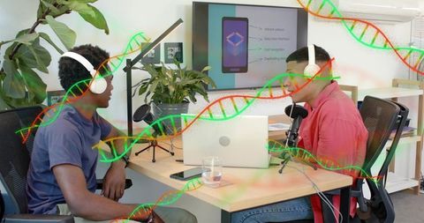 Recording podcast hosts wearing headphones at collaborative desk with laptop and microphones