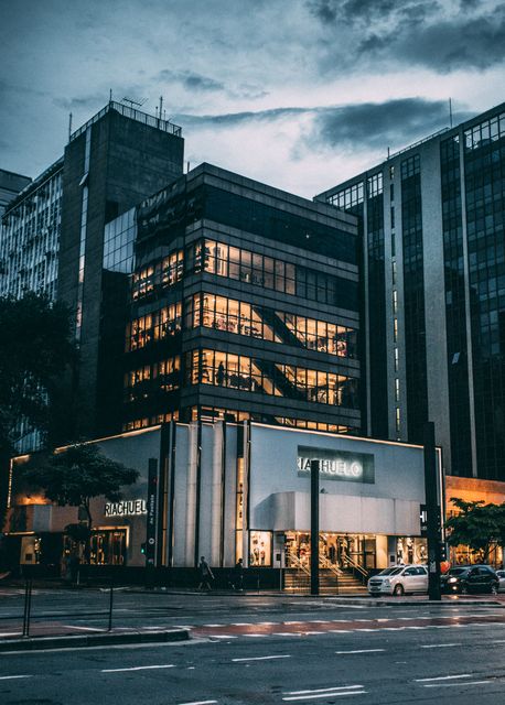 This image features a modern office building at dusk, prominently displaying its glass facade and warm interior lighting. The scene captures the urban life with a few vehicles and pedestrians on the street below. Perfect for use in architectural and real estate websites, city lifestyle magazines, and business brochures.