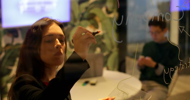 Businesswoman brainstorming on glass wall with colleague in background - Download Free Stock Images Pikwizard.com