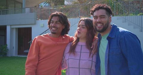 Young adults linking arms, laughing together in backyard near modern home terrace, sunny
