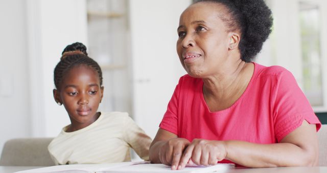 Elderly African American woman reading a Braille book at home while young girl intently watches. Ideal for use in educational content, literacy advocacy, inclusive education material, intergenerational bonding promotions, and family-based learning visuals.