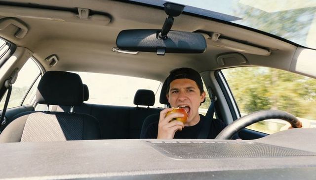 Driver biting apple while driving with backward cap and seatbelt, empty passenger seat