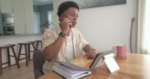 African american man working remotely on tablet and phone at kitchen table with notebook