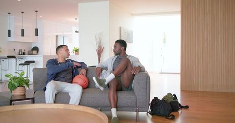 Multiracial young men relaxing on sofa holding basketball in minimalist living room