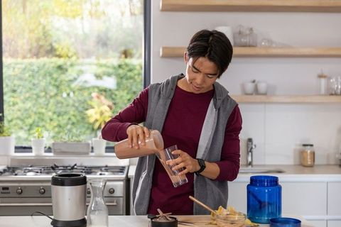Young asian man pouring chocolate smoothie from blender jar in modern sunlit kitchen for breakfast