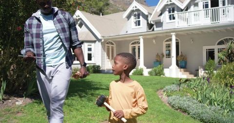 African american father and son walking on front lawn holding tablet and mallet, suburban porch