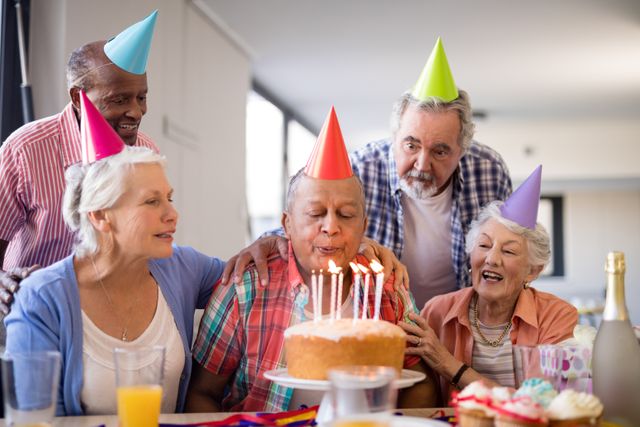 Senior friends celebrating a birthday in a nursing home. The group is gathered around a table with a birthday cake, wearing colorful party hats. The birthday person is blowing out candles while others smile and cheer. Perfect for illustrating themes of elderly care, community, friendship, and joyful moments in later life.
