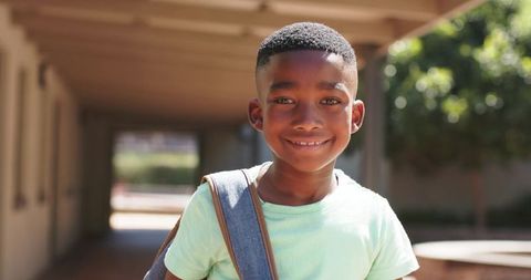 Smiling African schoolboy standing with backpack on sunlit campus walkway, portrait, casual