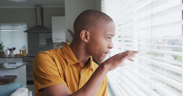 Man wearing yellow shirt is looking through window blinds in a modern kitchen. Ideal for concepts such as observation, curiosity, pondering, home life, anticipation, or waiting for something.