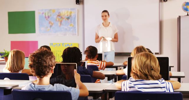 This image shows a teacher conducting a class with young students using tablets. This setup represents a modern classroom leveraging technology for interactive learning. Diverse students are seated and appear engaged with their tablets. This image is perfect for use in educational websites, technology in education articles, online learning platforms, and school promotional materials.