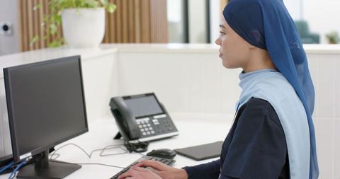 Female receptionist wearing blue hijab working at modern front desk computer in daylight office